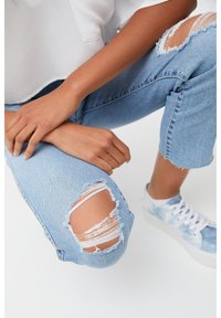 Jean déchiré bleu clair avec genoux effilochés, porté avec une chemise blanche et des baskets tie-dye bleues. La texture est en denim doux avec des accents usés.
