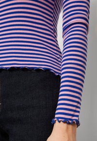 Zbliżenie na osobę ubraną w niebiesko-różową bluzkę z długim rękawem w paski, z rękawami o falbance, połączoną z ciemnoniebieskimi dżinsami.