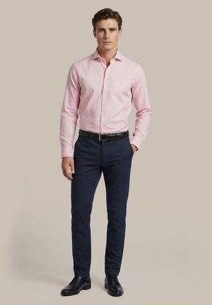 Mann trägt slim-fit hellrosa Hemd, dunkelblaue Hose, schwarzen Gürtel und schwarze Anzugschuhe, steht vor schlichtem beigen Hintergrund.
