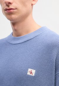 Sweater de malha tipo waffle azul claro com decote redondo, apresentando um pequeno patch do logotipo da Calvin Klein no canto inferior direito.