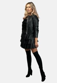 Czarna skórzana kurtka z teksturowanymi rękawami, warstwowa czarna spódnica z sztucznej skóry i czarne kozaki do kolan. Modelka pozuje w profilu.