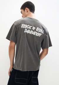Siva bomba majica iz bombaža z kratkimi rokavi in sproščenim krojem, z belim napisom "ROCK N' ROLL DAMNATION" na hrbtu.