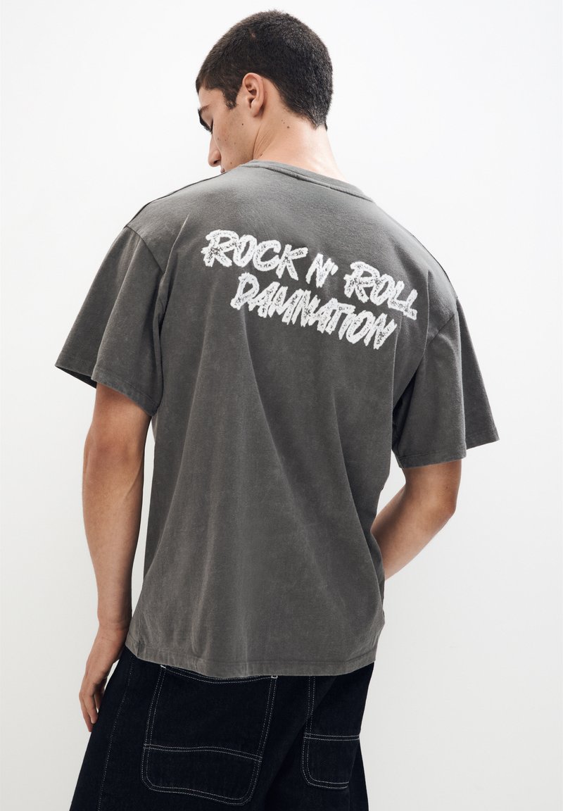 Siva bomba majica iz bombaža z kratkimi rokavi in sproščenim krojem, z belim napisom "ROCK N' ROLL DAMNATION" na hrbtu.