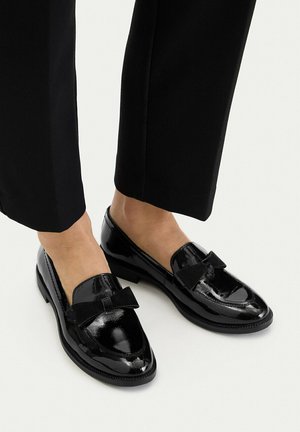 Mocasines de charol negro brillante con detalle de lazo negro, usados con pantalones negros recortados y pies descalzos.