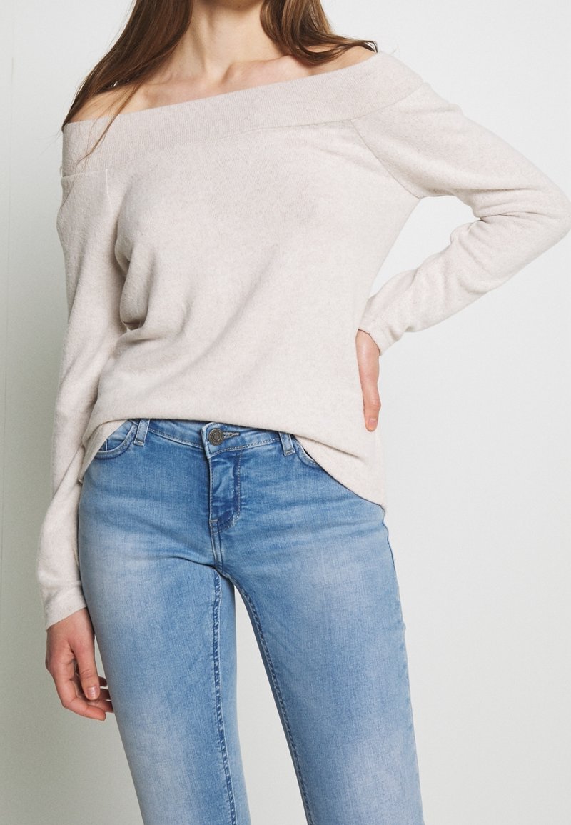 Femme portant un pull beige clair à épaules dénudées et un jean bleu délavé, debout, une main sur la hanche et l'autre détendue le long du corps.