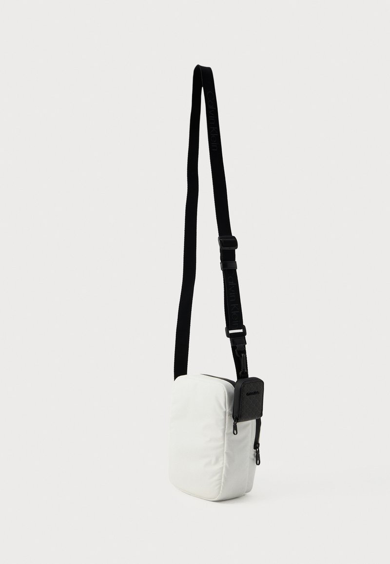 Bolso cruzado rectangular blanco con correa negra ajustable y pequeño bolsillo negro con cremallera adjunto.