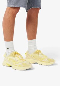 Person trägt hellgelbe Mesh-Sneaker mit weißen Sportsocken und grauen Jeansshorts, steht vor weißem Hintergrund.