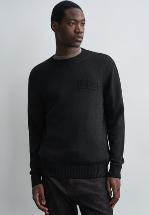 Fiatal férfi fekete kötött pulóvert visel, amelyen egy diszkrét mellkasi logó található, sötét nadrágban, egy sima világosszürke háttér előtt áll.