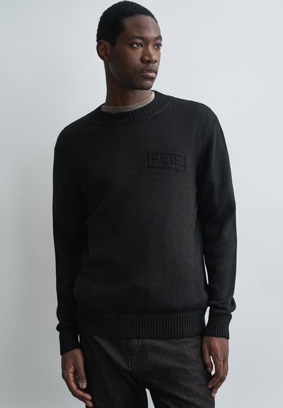 Giovane uomo con un maglione a maglia nero con un discreto logo sul petto, pantaloni scuri, in piedi davanti a uno sfondo grigio chiaro uniforme.