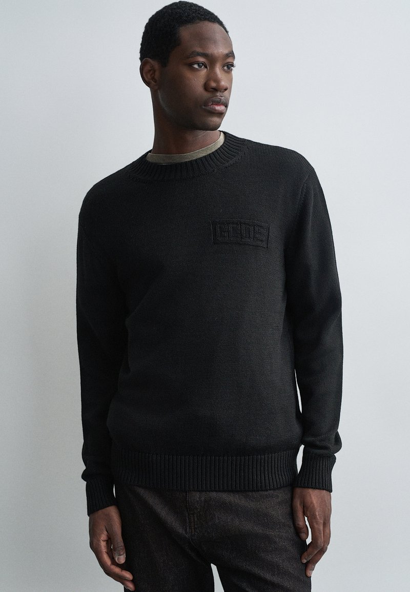 Jaunas vyras, vilkintis juodą megztinį su subtiliu logotipu krūtinėje, tamsias kelnes, stovi prieš šviesiai pilką fono.