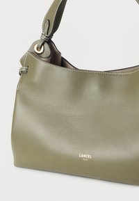 Δέρμα χρώματος ελιάς με λεία υφή, τσάντα χειρός με ένα λουρί ώμου και χρυσή λεπτομέρεια λογότυπου. Διαθέτει ραφές σε αντίθεση κατά μήκος των άκρων.