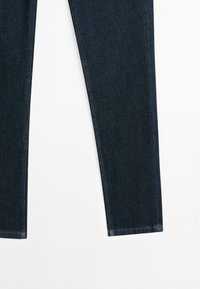 Jambes de jean en denim bleu foncé montrées à plat avec coutures latérales visibles et couture simple aux ourlets sur fond blanc.