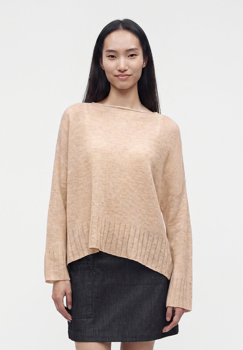 Femme aux longs cheveux foncés porte un pull beige ample à ourlet côtelé et une jupe noire, debout devant un fond clair uni.