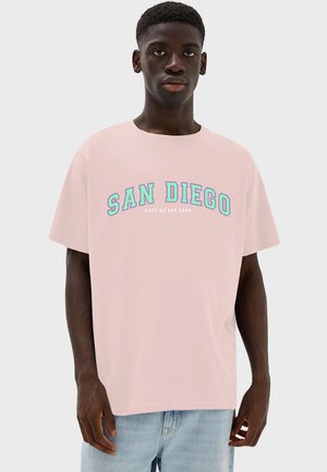 Světle růžové bavlněné tričko s nápisem "SAN DIEGO" v tučných tyrkysových písmenech. Klasický kulatý výstřih a krátké rukávy, nošeno s světle modrými džíny.