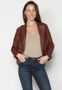 Giacca da moto in pelle marrone con tasche con zip, maniche lunghe e colletto classico, abbinata a jeans blu aderenti e una maglietta beige.