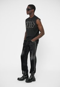 Svart ärmlös tröja med "KISS"-motiv och nitar, kombinerad med mörka jeans med en sidoseam prydd med silvernitar, avslutade med svarta stövlar.