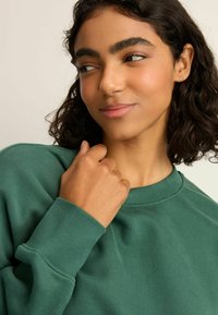 Groene katoenen sweatshirt met een ronde hals, geribbelde manchetten en een zachte textuur. Het model houdt de stof voorzichtig bij haar schouder.