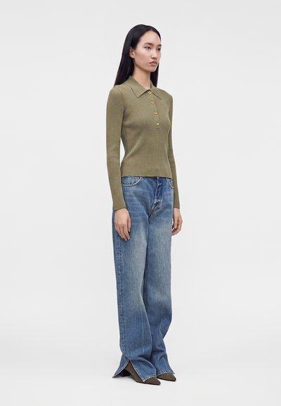 Femme aux longs cheveux noirs portant un haut côtelé à manches longues vert olive avec des boutons et un jean bleu à jambes larges avec ourlets fendus.