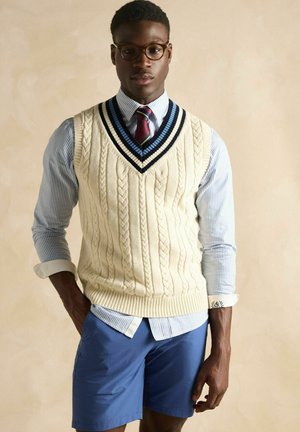 Cremfarbener Kabelstrick-Weste mit einem marineblauen gestreiften Ausschnitt über einem hellblau gestreiften Hemd, kombiniert mit blauen Shorts.