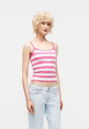 Ung person med kort krøllet blondt hår iført lyserød og hvidstribet tanktop og lyseblå jeans, der står mod en ensfarvet baggrund.