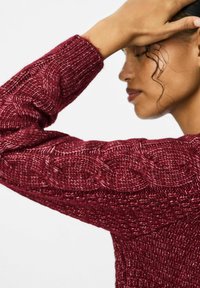 Sarkans adīts džemperis ar kabelīša adījumu uz piedurknēm un ribota aproce. Tekstūra ir masīva un ērta, ar pieguļošu dizainu.