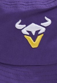 Gorra morada de tela, con un diseño de toro bordado en blanco y una forma de V amarilla en el centro. Superficie texturizada con costuras visibles.