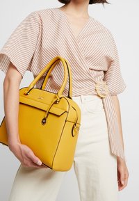 Żółta skórzana torebka o wyrazistym kształcie, z górnymi uchwytami i zapięciem na klamrę z przodu. Posiada fakturowane wykończenie oraz czarne akcenty dla kontrastu.