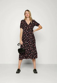 Mulher em pé, vestindo um vestido midi preto com padrão floral e sapatos pretos tipo slip-on, segurando uma pequena bolsa preta contra um fundo claro e liso.