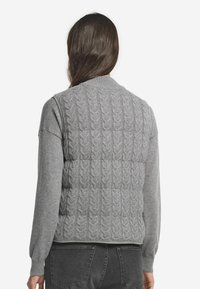 Kvinna med långt mörkt hår bär en grå kabelstickad ärmlös väst över en långärmad grå tröja och svarta jeans, vänd bort.