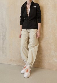 Czarna polarowa kurtka na zamek, jasnoróżowa koszulka z grafiką, beżowe spodnie cargo z kieszeniami oraz białe trampki z pomarańczowymi sznurowadłami i szarymi akcentami.