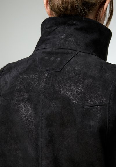 Personne portant une veste en daim noire avec un col relevé, montrant des détails de couture et un petit logo embossé sur le haut du dos.