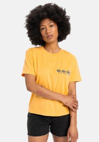Yellow cotton t-shirt with short sleeves and a rounded neckline. Features a graphic design of three birds on the chest. Worn with black shorts.