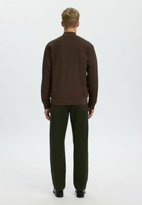 Brūna zip-up jaka ar teksturētu virsmu, ribota apakšmalas un taisnu apakšdaļu, kombinēta ar zaļām platām biksēm un melniem apaviem.