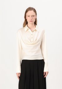 Blusa a maniche lunghe color crema con scollo a cappuccio e dettaglio dei bottoni sul davanti, realizzata in un tessuto liscio simile alla seta.