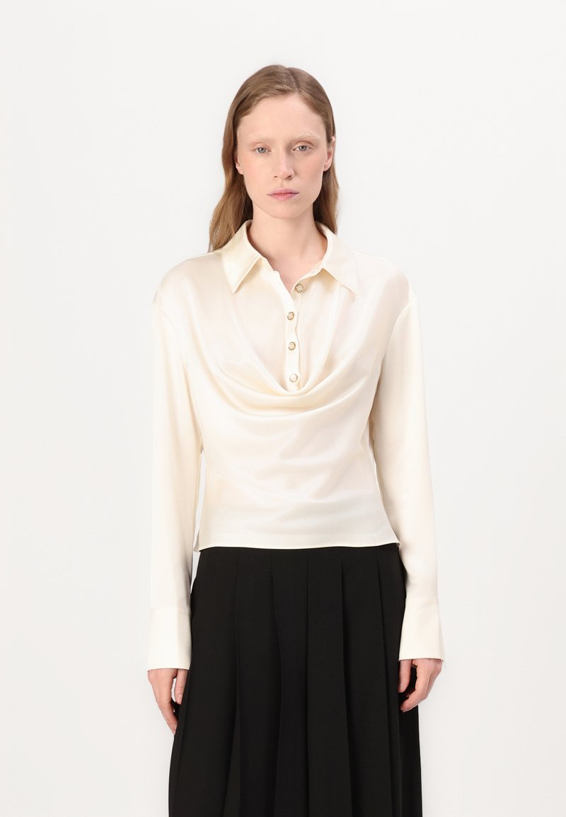 Blusa a maniche lunghe color crema con scollo a cappuccio e dettaglio dei bottoni sul davanti, realizzata in un tessuto liscio simile alla seta.