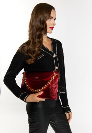 Porte-monnaie en paillettes rouges avec une chaîne dorée, légèrement arrondi en haut et ayant une texture brillante, tenu contre un pull noir et un pantalon en cuir.