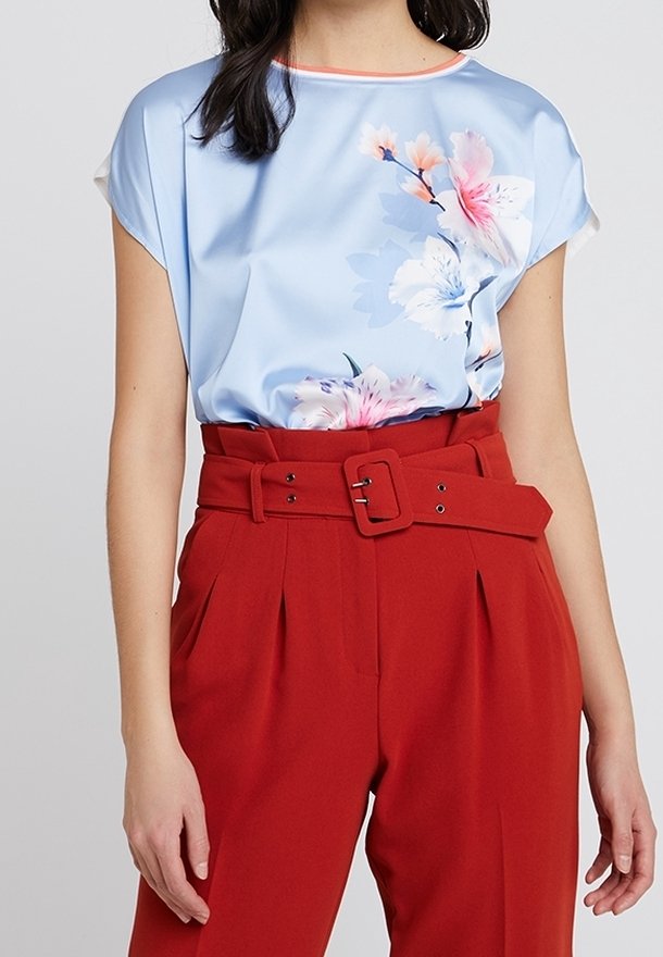 Woman wearing a light blue satin blouse with pink and white floral print, tucked into high-waisted red trousers with a wide belt and buckle.