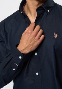 Granatowa koszula zapinana na guziki z usztywnianym kołnierzykiem, białymi guzikami i wyszywanym logo. Posiada kontrastujący wewnętrzny detal kołnierzyka.