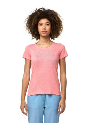 Une femme aux cheveux bouclés se tient devant un fond blanc, vêtue d’un tee-shirt rose Leone et d’un pantalon bleu clair.