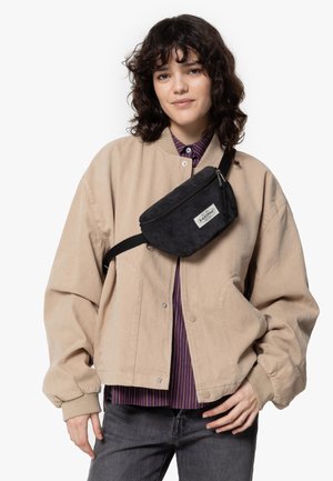 Junge Frau mit lockigem, dunklem Haar, die eine beigefarbene Jacke, ein gestreiftes Shirt, dunkle Jeans und eine schwarze Umhängetasche trägt, vor einem schlichten weißen Hintergrund.