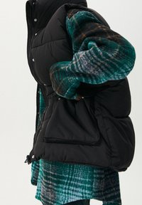 Černá prošívaná vesta se zapínáním na zip a druky, vrstvená přes zeleno-modrou kostkovanou košili, zdůrazňující texturované materiály.