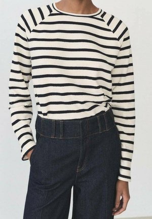 Personne portant une chemise à manches longues rayée crème et noire rentrée dans un jean bleu foncé taille haute, main dans la poche.