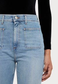 Jeans de tiro alto de color azul claro, confeccionados en denim, con cierre de botón, pasadores para cinturón y dos bolsillos traseros con costuras visibles.