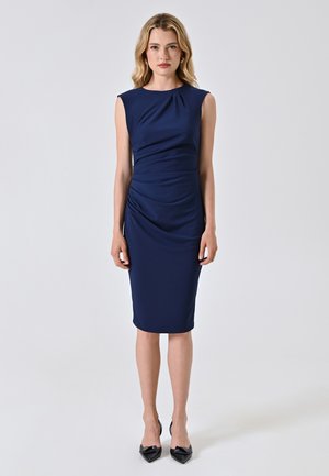Abito fitted blu navy con maniche a cappuccio, caratterizzato da dettagli arricciati in vita e un orlo a lunghezza ginocchio. Tessuto liscio e elasticizzato. Tacchi neri a punta.