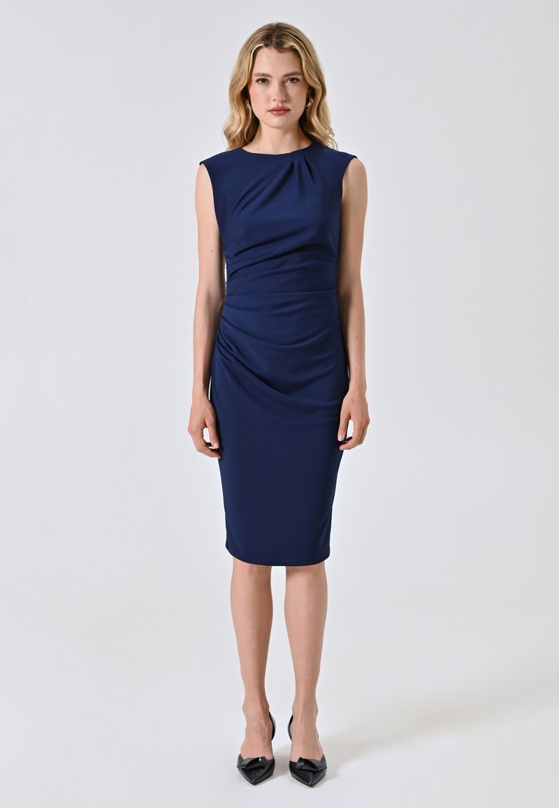 Robe ajustée marine avec des manches à capuchon, présentant des détails froncés à la taille et une longueur jusqu'au genou. Tissu lisse et extensible. Escarpins noirs à talons pointus.