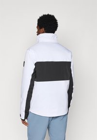 Λευκό μπουφάν με μια μαύρη οριζόντια ρίγα στη πλάτη. Διαθέτει ψηλό γιακά και αντίθετες μαύρες πλευρικές πάνελ. Λείος υφασμάτινος ιστός.