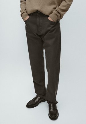 Person iført mørkebrune bukser med afslappet pasform, sorte loafers og en lysebrun sweater med hænderne i lommerne.