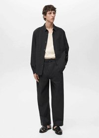 Schwarze Button-Down-Bluse über einem beigen Henley, kombiniert mit lockeren schwarzen Hosen und schwarzen Sandalen. Die Bluse hat ein strukturiertes Design und eine Brusttasche.