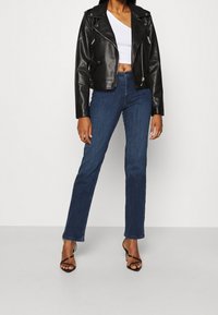 Chaqueta de cuero negro de moto recortada con cremalleras plateadas, combinada con unos vaqueros de mezclilla azul de pierna recta. Sandalias de tacón alto negras con tiras completan el look.