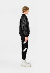 Chaqueta bomber de cuero negro con puños acanalados, combinada con pantalones jogger negros que tienen gráficos de plumas blancas y botas de combate blancas.